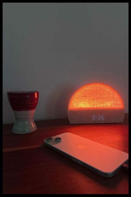 The Hatch Restore 2, displaying the time and lit-up in an orange glow, sitting atop the writer's wooden dresser. There is also an iPhone charging in front of the clock, and to the clock's left is a small clay vase.