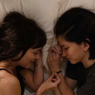 Photo of two women laying in bed, looking at each other. 