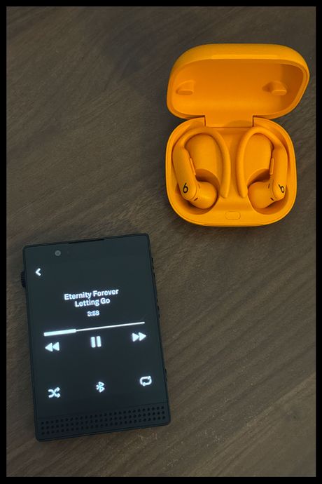 The Light Phone III with the song “Letting Go” by Eternity Forever on the screen, and a pair of orange Powerbeats Pro earbuds next to the phone.