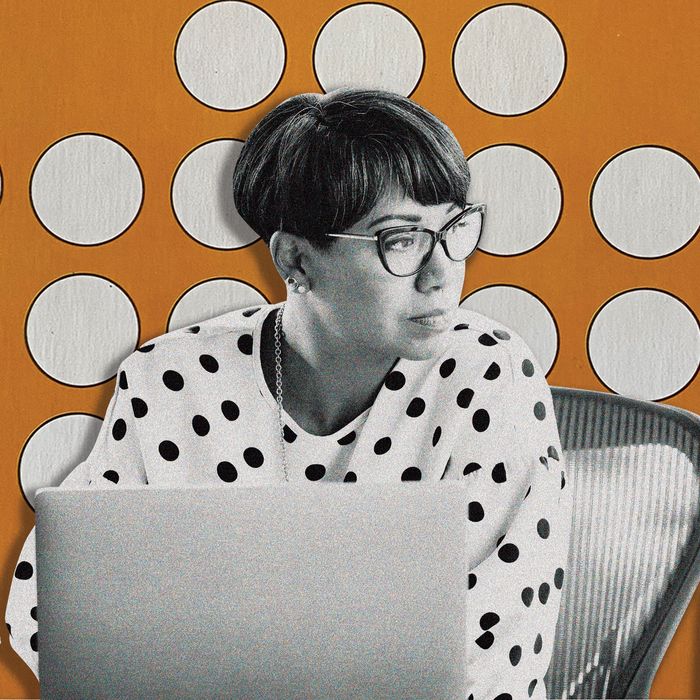 A woman who has short, cropped black hair and is wearing cat-eye glasses and a black-and-white polka dot top looks off to her left. She is sitting behind a laptop and there is a coffee cup on her desk. 