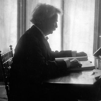 Profile of Mark Twain seated at a desk, reading a large book, 1906. 