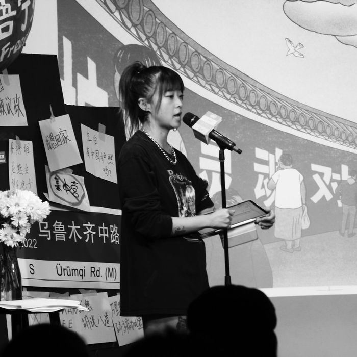 A young Chinese woman wearing a black tee shirt and black hot pants stands at a microphone.