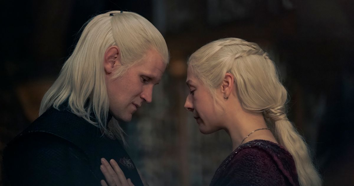 A man and a woman, both with bright blonde hair, stand face to face. The woman has her hands on the man's chest, looking there. The man is also looking down. They both look solemn.