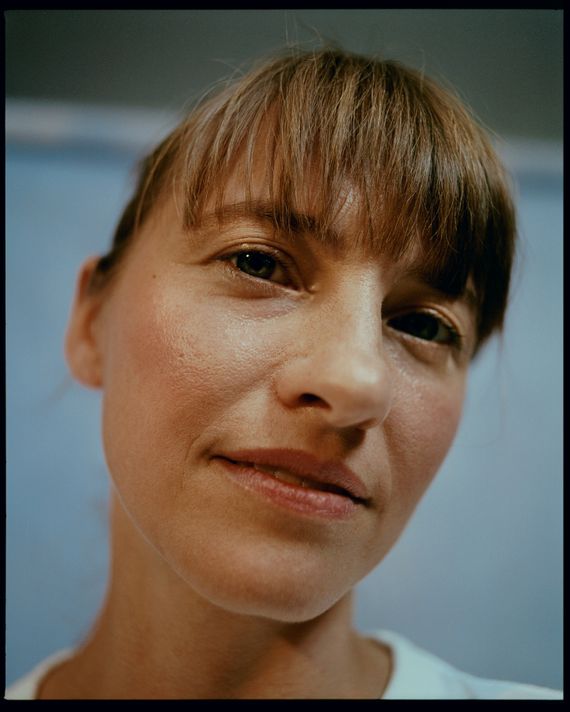 A woman looks at the camera for a close-up portrait. She has brunette hair that's pulled back and bangs covering her forehead. She's wearing a white T-shirt, and her face is turned upward to the camera slightly, her blue eyes making contact with the lens.