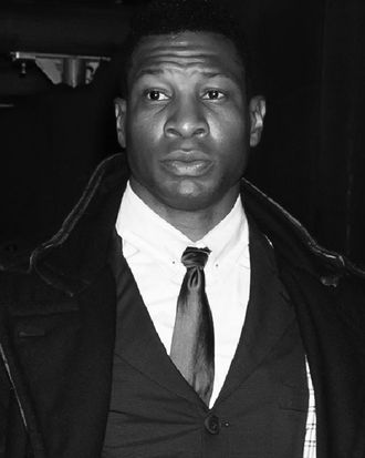 A black man wearing a white collared shirt, a dark tie, a dark waistcoat, and a dark overcoat looks past the camera with his eyebrows slightly raised. 