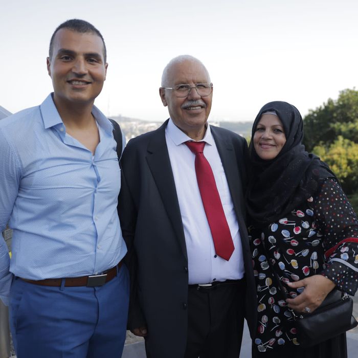 A man in a blue dress shirt, a shorter man in a suit, and a woman wearing a hijab smile for a family photo.