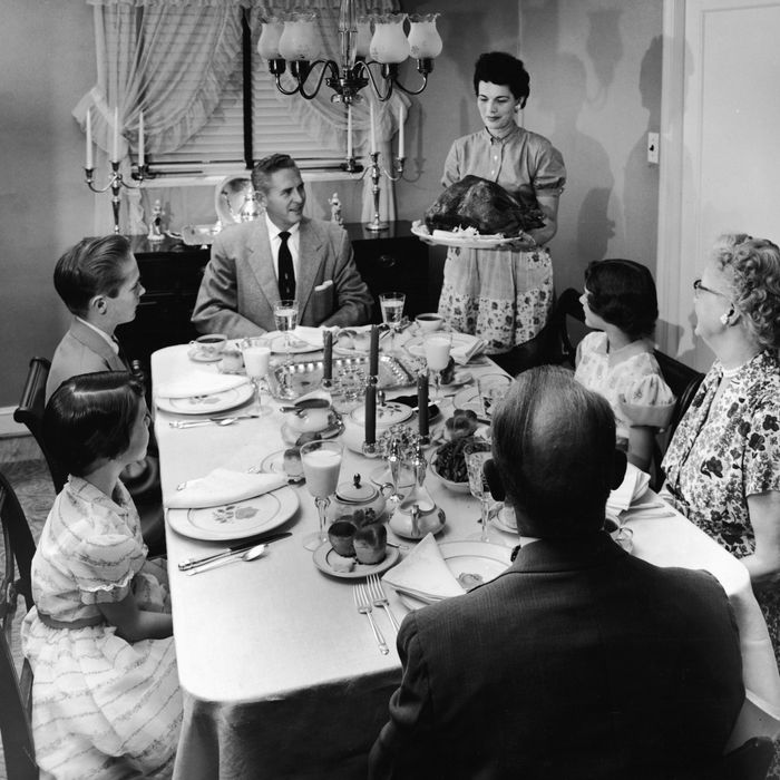 A family sits at a formally prepared dinner table, looking eager as a woman brings a roast turkey to the table on a platter, 1950s.