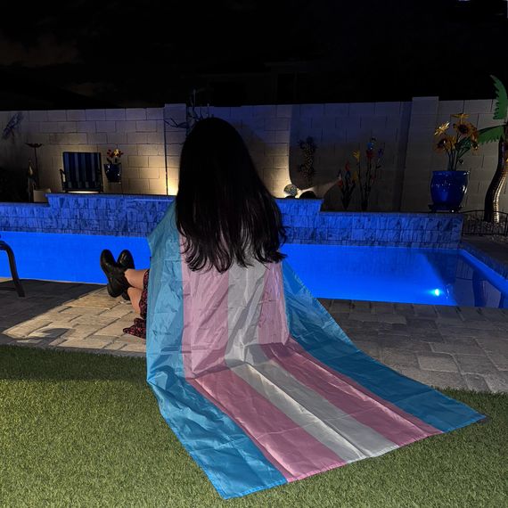A girl sits at the edge of a swimming pool, wearing a trans flag as a cape.