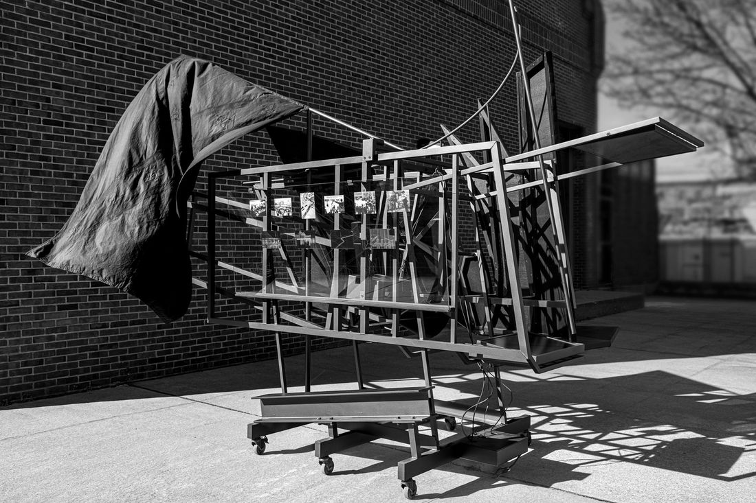 A black aluminum and wooden structure on wheels with video screens and photos mounted to it