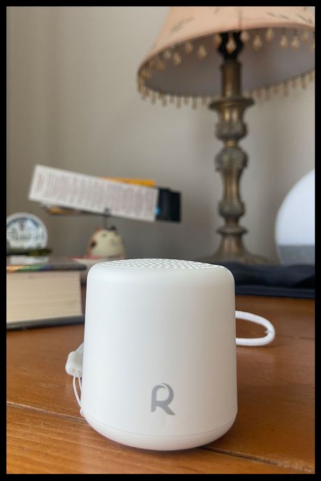A small white sound machine on a bedside table, with a lamp and book in the background.