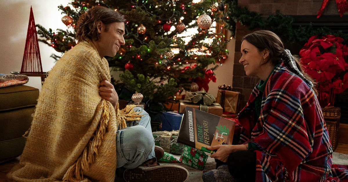 The male and female lead from Hot Frosty are sitting on the floor facing each other, smiling, and wearing blankets around their shoulders. They are opening Christmas presents.
