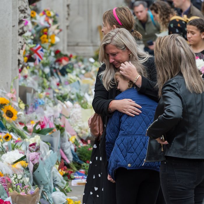 A woman with long blonde hair hugs a young girl whose hair is in braids to her chest. They're looking through tears at a makeshift memorial of flowers for the late Queen Elizabeth laid at the gates of Buckingham Palace. 