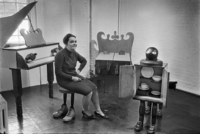 Kate Millett sits on a stool with legs that look like stockinged feet. She's surrounded by sculptural furniture like a non-playable piano with two wooden fists hovering above the keyboard, a cabinet that looks like a person, and a bed with two wood figures resting on it.